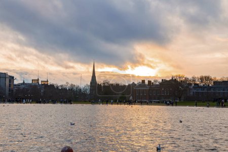 Téléchargez les photos : Vue panoramique sur le magnifique lac et le jardin avec l'église St Mary Abbots et le palais de Kensington en arrière-plan. Journée nuageuse au coucher du soleil à Londres - en image libre de droit
