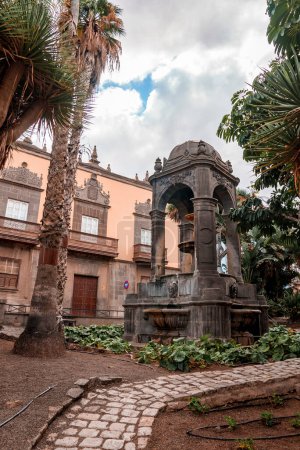 Foto de Low angle view of fountain with medieval architectural structure with trees and building at Plaza del Espiritu Santo in Vegueta on a cloudy day at Las Palmas, Gran Canaria, Spain - Imagen libre de derechos