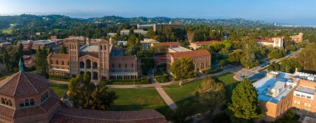 Foto de Vista aérea del campus de UCLA con torre gótica, edificios de ladrillo rojo, césped verde y caminos en medio de un paisaje montañoso cubierto de árboles con luz suave y dorada. - Imagen libre de derechos