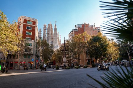 Photo for Daytime Barcelona street scene with La Sagrada Familias spires in the background, treelined avenues, and a mix of parked vehicles under a clear blue sky. - Royalty Free Image