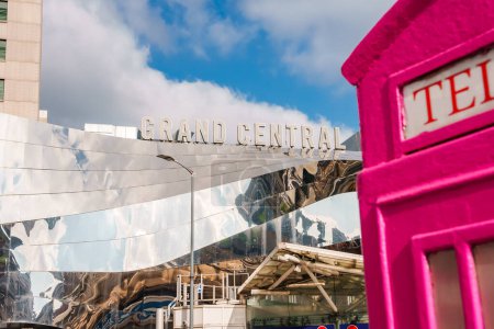 Moderne Architektur des Grand Central Birmingham mit metallischen Buchstaben, reflektierenden Oberflächen und einer leuchtend rosafarbenen Telefonzelle vor einem teilweise bewölkten Himmel, die Stadtentwicklung und britisches Erbe präsentiert.