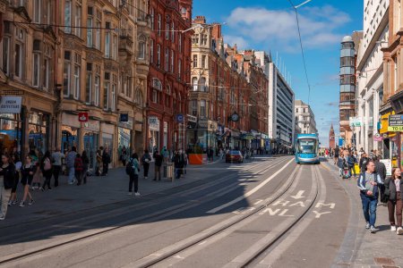 Foto de Un moderno tranvía azul se desliza a través de Birmingham, Reino Unido, bajo un cielo despejado, flanqueado por una mezcla de edificios históricos y elegantes, con aceras bulliciosas que insinúan una animada vida en la ciudad. - Imagen libre de derechos