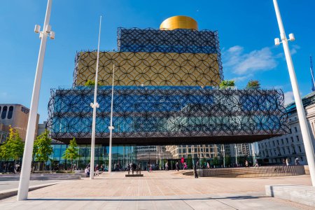 Téléchargez les photos : Une journée ensoleillée met en valeur la Bibliothèque de Birmingham, au Royaume-Uni, avec sa façade métallique à motifs et sa rotonde dorée sur le toit. Les gens apprécient la place bordée de mâts de drapeau, mettant en évidence le point de repère culturel des villes. - en image libre de droit