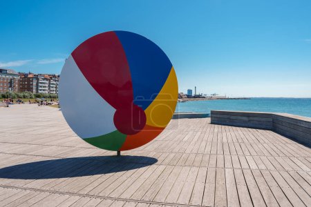 Photo for A vibrant circular sculpture with six colored segments stands on a wooden boardwalk by the sea, with a sandy beach and urban backdrop, likely in Helsingborg. - Royalty Free Image