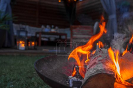 Evening ambiance with a lively fire in a metal pit, casting a warm glow in a garden with a wooden cabin or bar in the background, possibly in Copenhagen.