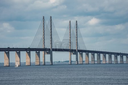 The Oresund Bridge, a marvel connecting Malmo, Sweden, with Copenhagen, Denmark, stands out with its cablestayed design and towering pylons against an overcast sky, with a sailboat below.