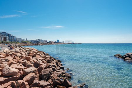 Photo for A serene coastal landscape with a rocky shoreline and calm waters under a clear blue sky. Modern buildings line the coast, suggesting an urban beachfront in Helsingborg. - Royalty Free Image