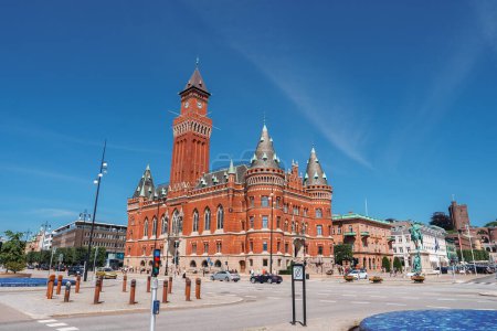 Téléchargez les photos : Un bâtiment en brique rouge ensoleillé avec des éléments néoromans et gothiques, doté d'une tour et de fenêtres cintrées, se dresse sur une place calme à Helsingborg. - en image libre de droit