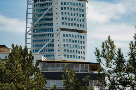 Foto de Un rascacielos moderno, el Turning Torso en Malmo, Suecia, gira hacia el cielo con una fachada blanca y un patrón de ventanas, enmarcado por siempreverdes en un día claro con cielos azules. - Imagen libre de derechos