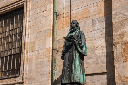Téléchargez les photos : Une statue en bronze d'une figure historique saisissant un livre se dresse devant un bâtiment classique, peut-être à Copenhague, sous un ciel clair aux ombres douces accentuant sa forme. - en image libre de droit