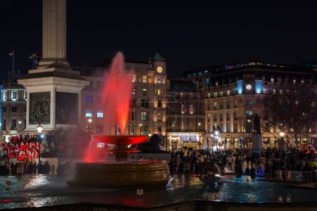 Téléchargez les photos : Une soirée festive à Trafalgar Square, Londres, avec une fontaine rougeoyante rayonnante, des foules animées, et peut-être un chœur en rouge, près de la colonne Nelsons partiellement visible. - en image libre de droit