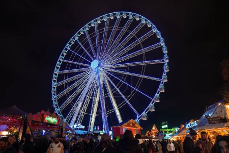 Photo for A lively London Winter Wonderland scene with a static, brightly lit Ferris wheel, bustling crowds in winter attire, and neonlit stalls offering festive attractions. - Royalty Free Image