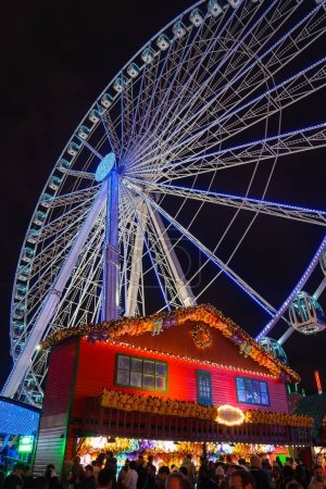 Téléchargez les photos : Une scène festive de nuit londonienne avec une grande roue rayonnante et un chalet décoré de lumières de Noël, entouré d'une foule animée, capturant l'esprit de vacances. - en image libre de droit