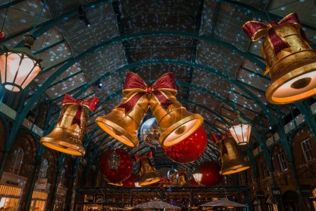 Photo for A festive London Covent Garden setting with a vaulted glass ceiling, adorned with large golden bells and red baubles, exudes Christmas cheer in a warm, ambient light. - Royalty Free Image
