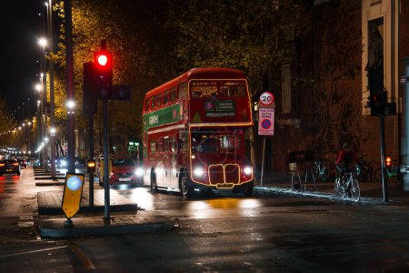 Téléchargez les photos : Une scène londonienne festive la nuit avec un bus double étage rouge décoré offrant des lumières de jardin et des tours de thé de l'après-midi sous les lampadaires sur une route mouillée, avec un cycliste et la circulation en mouvement. - en image libre de droit