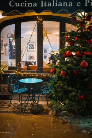 Téléchargez les photos : Un charmant restaurant italien à Londres, le Cucina Italiana e Pizza, avec un cadre extérieur festif doté d'un ensemble bistro bleu, d'un arbre de Noël décoré et d'un éclairage chaleureux reflétant un trottoir humide.. - en image libre de droit