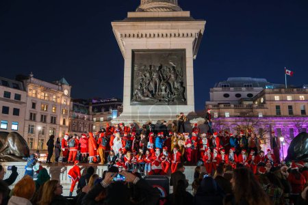 Foto de Una animada reunión de personas en trajes de Santa celebra la Navidad en los escalones de un monumento clásico en Londres, Reino Unido, bajo un cielo nocturno azul oscuro, con espectadores capturando el momento alegre. - Imagen libre de derechos