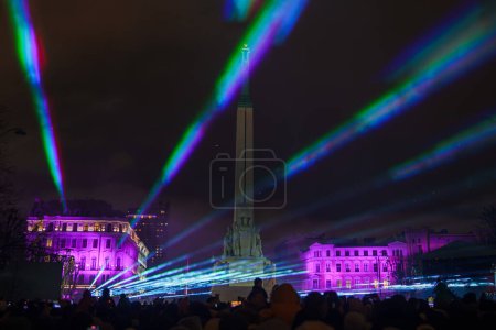 Téléchargez les photos : Célébration nocturne à Riga, Lettonie, avec des spectateurs en silhouette admirant le monument de la liberté illuminé au milieu d'un spectacle lumineux animé de poutres vertes, bleues et roses, flanqué de bâtiments classiques. - en image libre de droit