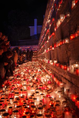 Téléchargez les photos : Une scène solennelle de commémoration avec des bougies dans des bocaux en verre bordant un mur de briques, alors que les gens s'agenouillent en réflexion près d'une croix, probablement pendant la fête de l'indépendance de Lettonie. - en image libre de droit
