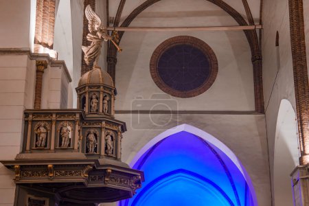 Téléchargez les photos : Une chaire en bois finement faite avec des détails en or et des figures sacrées dans une cathédrale du dôme de Riga, éclairée par un ange suspendu et des lumières bleues, probablement en Lettonie pour le jour de l'indépendance. - en image libre de droit