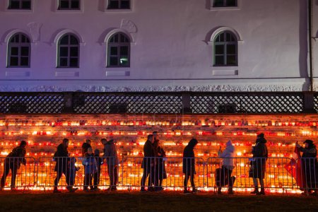 Foto de Una noche serena en Letonia con individuos reunidos para un evento a la luz de las velas, reflexionando sobre el Día de la Independencia. El cálido resplandor de las velas en las escaleras contrasta con el crepúsculo y un modesto edificio blanco. - Imagen libre de derechos