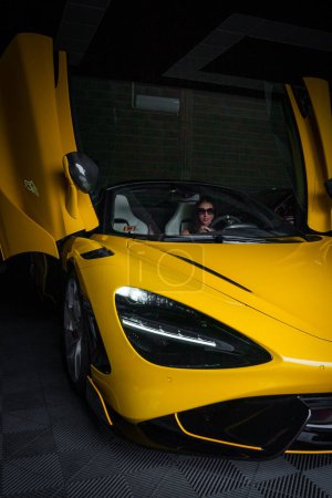Téléchargez les photos : Une McLaren 765LT jaune brillant avec portes dièdres ouvertes, assise dans un garage d'accordage faiblement éclairé, présentant son design sportif et ses lignes aérodynamiques, avec une personne assise à l'intérieur. - en image libre de droit
