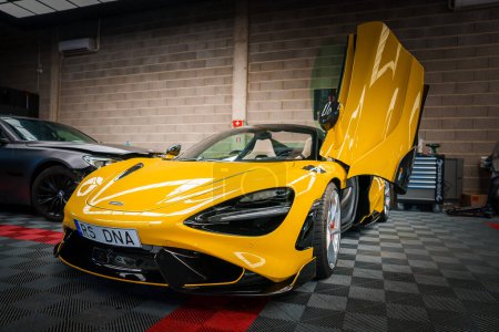 Photo for A vibrant yellow McLaren 765LT with upwardopening dihedral doors is displayed in a tuning garage, featuring black aerodynamic accents and multispoke wheels against a checkered floor. - Royalty Free Image