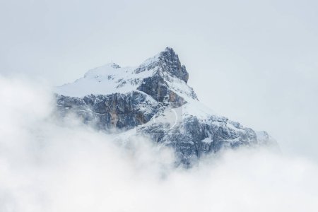 A snowcovered mountain peak rises above clouds in Engelberg, Switzerland, with a serene mix of whites and grays, highlighting the rugged, challenging terrain.