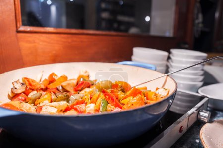 Closeup of a blue frying pan with colorful stirfried vegetables, including bell peppers and onions, cooked to a tender sheen, ready to serve in an Engelberg luxury hotel kitchen.