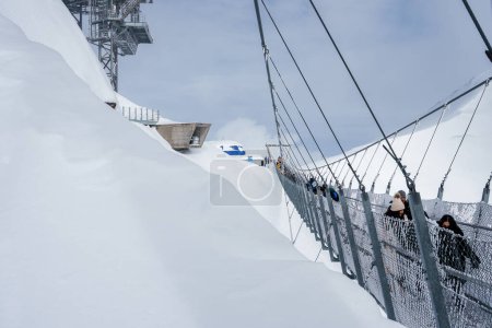 Photo for Suspension bridge at Engelberg ski resort in the Swiss Alps, with visitors crossing amidst heavy snow and a ski lift structure visible, under an overcast sky. - Royalty Free Image