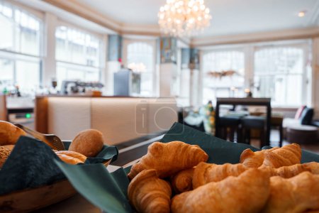 Photo for Closeup of freshly baked bread and croissants in a basket with a dark green napkin, set in a luxury hotels dining area with warm lighting, large windows, and elegant decor at Engelberg ski resort. - Royalty Free Image