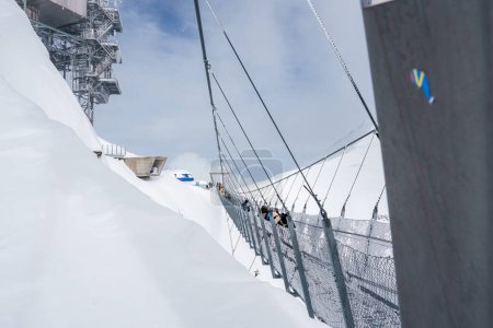 A serene, snowy scene at Engelberg, Switzerland, featuring a safe, metal cable bridge with mesh fencing, used by visitors in winter gear, with ski resort infrastructure in the background.
