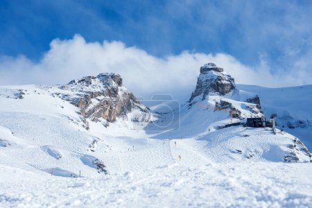 Téléchargez les photos : Une station de ski tranquille d'Engelberg avec des pistes enneigées, des pistes et un chalet alpin au bord des Alpes suisses. Les sommets ensoleillés et le ciel bleu clair ajoutent à l'atmosphère sereine et opulente. - en image libre de droit