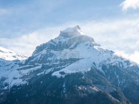 Temprano en la mañana o al final de la tarde la luz baña un pico nevado de la montaña Engelberg, con afloramientos rocosos y laderas boscosas bajo un cielo azul pálido con nubes tenues.