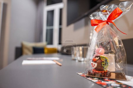 Primer plano de un caprichoso regalo de chocolate envuelto en celofán con una cinta roja, con detalles coloridos sobre una base estampada, frente a un interior borroso habitaciones modernas en Engelberg.