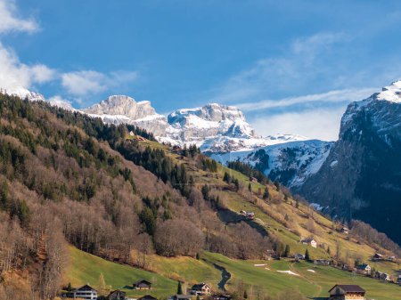 Scenic Engelberg, Switzerland, showcases a blend of greenery and snow with alpine houses dotting the slopes, while the majestic Swiss Alps rise in the background under a clear blue sky.