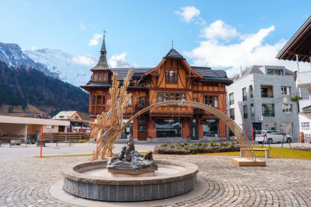 Photo for A serene Engelberg scene with a fountain sculpture, a Swissstyle building with a gabled roof, and contrasting modern architecture, set against snowcapped mountains and a clear sky. - Royalty Free Image