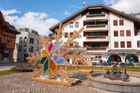 Photo for A vibrant snowflake sculpture with stained glass center adorns a sunny Engelberg town square, with a traditional fountain, Swiss flag, alpine architecture, and a relaxed outdoor cafe scene. - Royalty Free Image