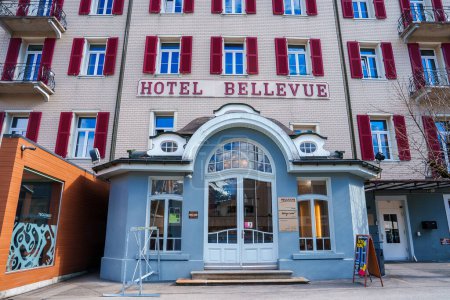 Foto de Clásico hotel de estilo europeo Bellevue en Engelberg con una fachada de color crema, persianas rojas y una entrada de arco azul, que ofrece un ambiente acogedor para los huéspedes del complejo de montaña. - Imagen libre de derechos