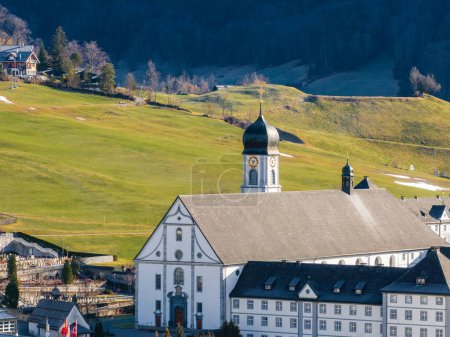 Capturando la esencia de Engelberg, Suiza, esta serena imagen cuenta con un hotel alpino blanco y una iglesia con una torre coronada de oro con un telón de fondo de verdes pendientes marrones y colinas boscosas.