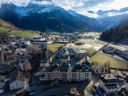 Photo for A serene aerial view of Engelberg in the Swiss Alps, showcasing chaletstyle buildings and a grand central hotel amid snowcapped peaks and green valleys. - Royalty Free Image