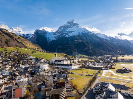 Photo for A scenic view of Engelberg in the Swiss Alps, showcasing a blend of traditional and modern architecture, a church spire, and snowcapped mountains under a clear sky. - Royalty Free Image