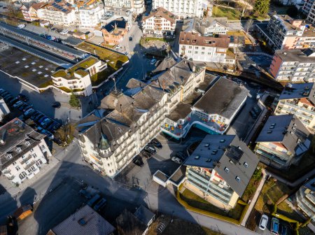 Aerial view of Engelberg, Switzerland, showcasing a large hotel with a white facade, colorful leisure buildings, diverse architecture, and a train station, under clear skies.