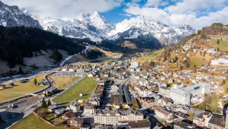 Téléchargez les photos : Vue aérienne de la station Engelberg dans les Alpes suisses, qui montre des montagnes enneigées, des forêts alpines et un mélange de chalets et de bâtiments modernes, avec un hôtel bien en vue et des parkings achalandés. - en image libre de droit