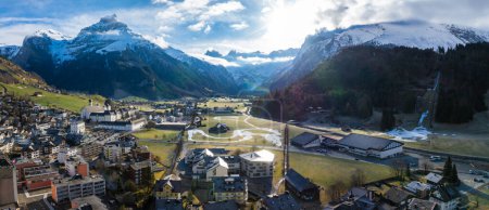 Panoramic view of Engelberg, Switzerland, showcasing its alpine architecture, a valley with a reflective river, and snowcapped mountains under a clear sky with a rainbow effect.