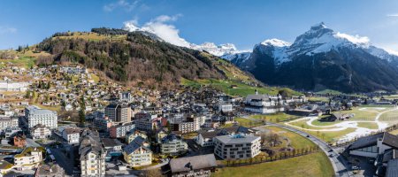 Vue panoramique de la station de ski Engelberg en Suisse, présentant un mélange de chalets et de bâtiments modernes au milieu de champs verts, de neige résiduelle et de montagnes alpines imposantes.