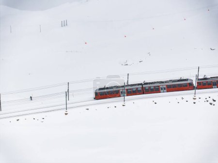 Aerial shot a bright red train glides through snowy Zermatt, beside a lone figure walking near the tracks. Sparse ski signs and red flags dot the serene landscape.