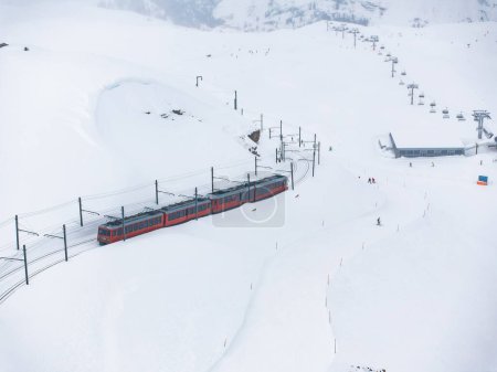Aerial shot of busy Zermatt ski resort, with a red train on snowy tracks and a ski lift going up the mountain. Small skiers dot the scene, enjoying winter sports on a chilly, cloudy day.