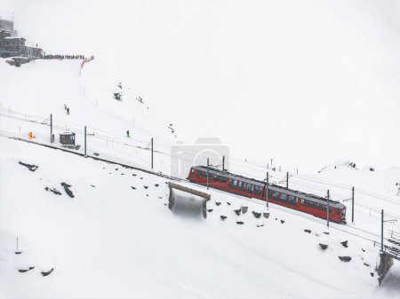 Aerial view of a vibrant red train emerging from a tunnel in Zermatt, Switzerland, against a snowy landscape. Skiers gather atop a slope, with mountains shrouded in fog or snowfall.
