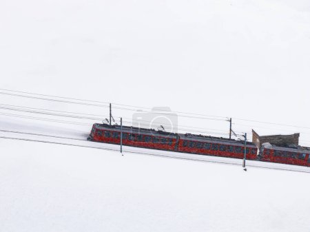Aerial shot of a red cogwheel train in Zermatt, Switzerland, gliding through snow. Its tracks stand out, highlighting its lone trip in a famed alpine spot.